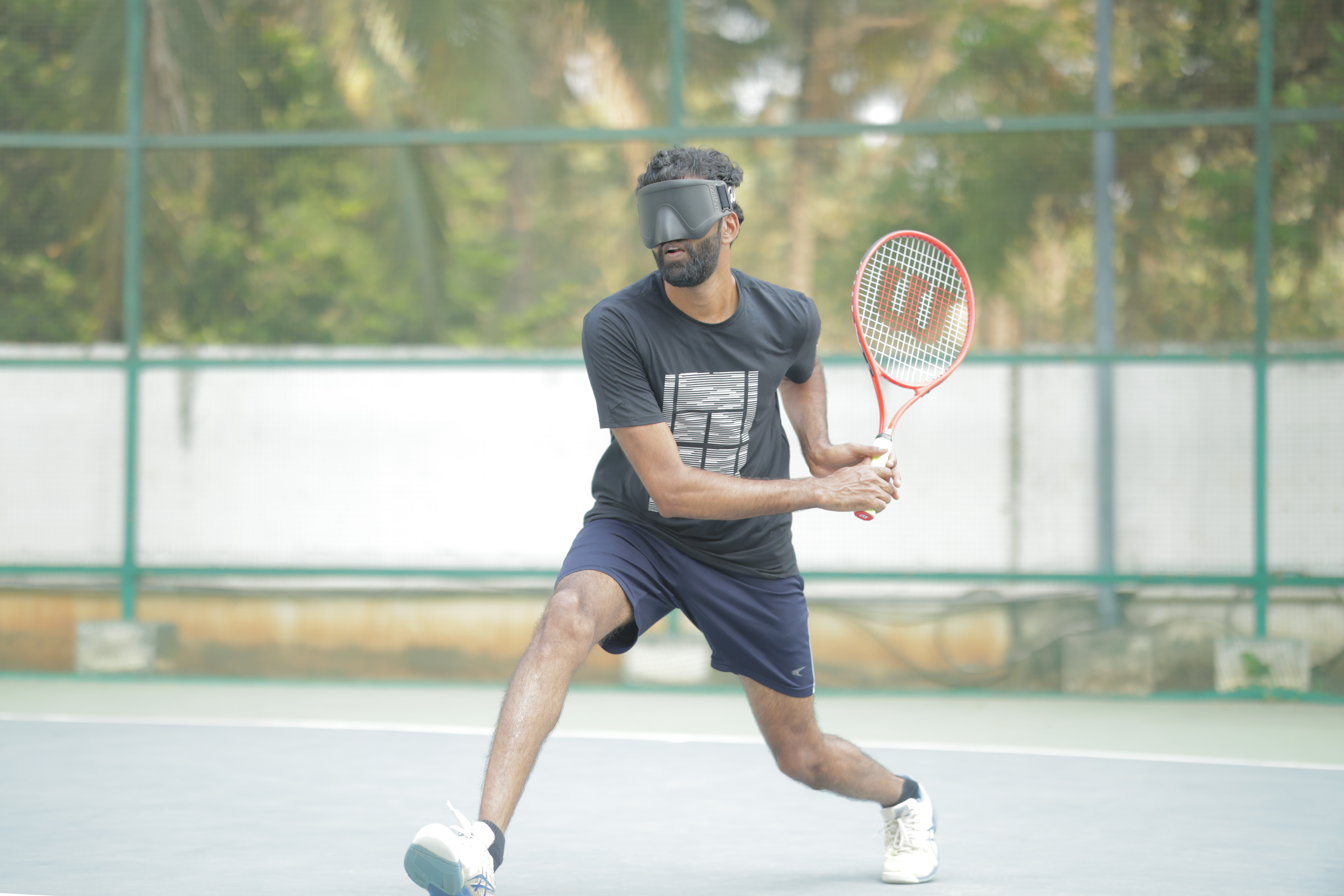 Nibin Mathew mid-swing on a tennis court, racquet connecting with the ball during a blind tennis match.